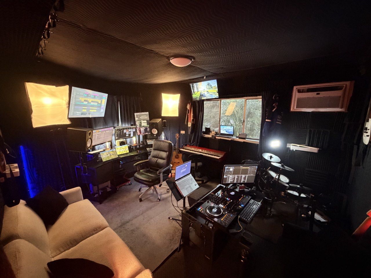 Clean studio view from couch — DAW monitors, softbox lights, DJ controller, drums